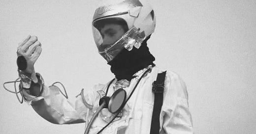 Gray-toned photo of a figure in a foil helmet and wired white suit, one hand raised in fog